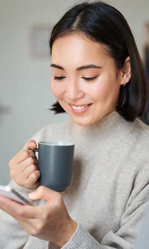 Portrait of smiling asian girl checking her news feed on smartphone and drinking coffee, sitting on sofa at home, browsing on mobile phone, reading.