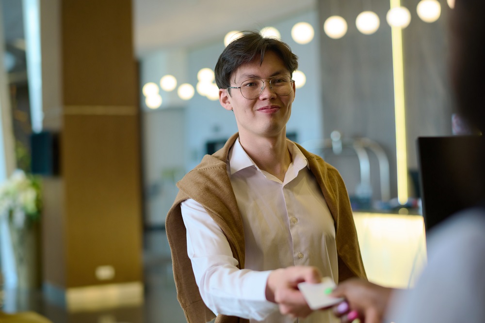 Happy young tourist in smart casualwear passing keycard to receptionist Asian business traveler checking in to hotel