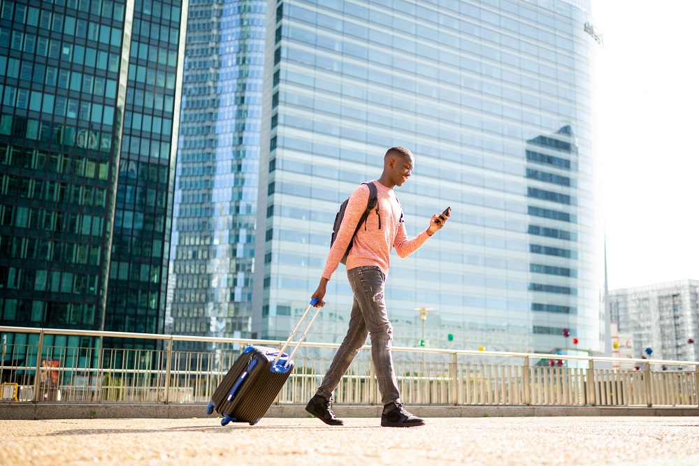 Full length side of young black man walking with mobile phone and pulling suitcase in city Happy business traveler walking with suitcase in city