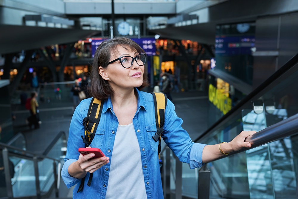 Woman on escalator stairs in building of city transport station with smartphone Frau auf der Rolltreppe mit Handy in der Hand, Geschäftsreisetrends