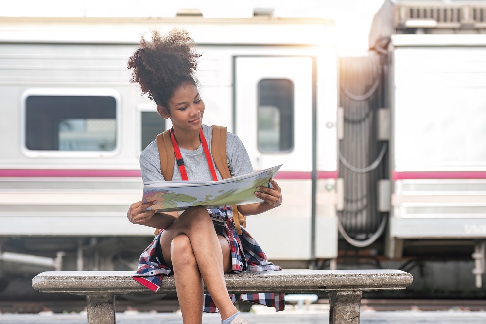 Young woman female smiling traveler with back pack looking to map while waiting for the train at train station. High quality photo Frau sitzt auf einer Bank im Bahnhof und hält eine Karte, während sie auf einer Bleisure-Reise ist