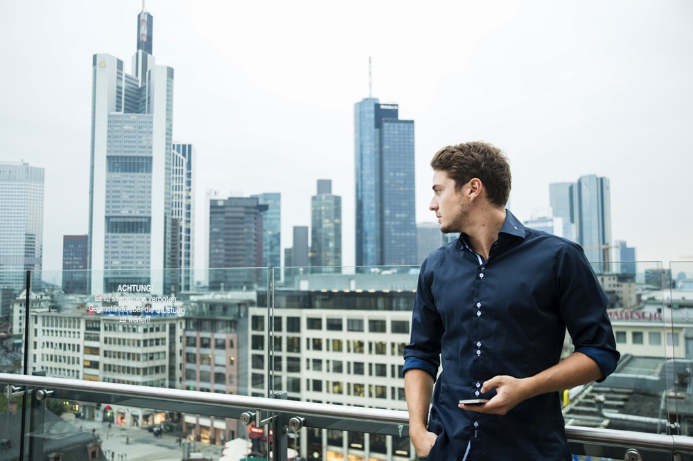 Germany, Hesse, Frankfurt, young man with his smartphone standing in front of the skyline Mann vor der Skyline von Frankfurt mit Smartphone in der Hand blickt in die Ferne