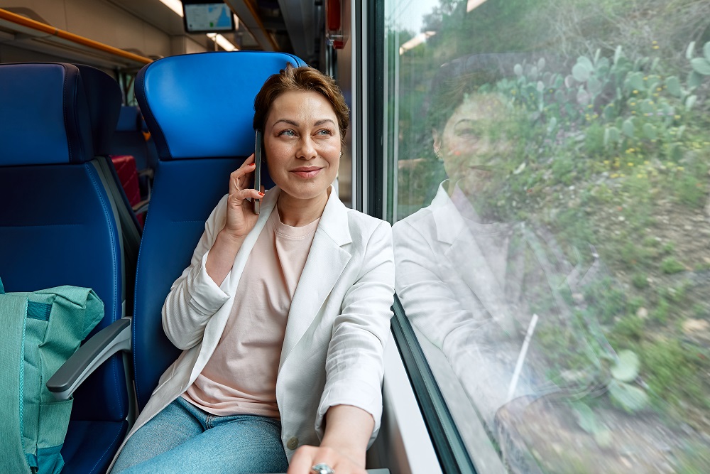 Smiling businesswoman freelance blogger using mobile phone in train. Pretty woman tourist traveling by train sitting near window and talking on smartphone. Geschäftsreisen mit der Bahn