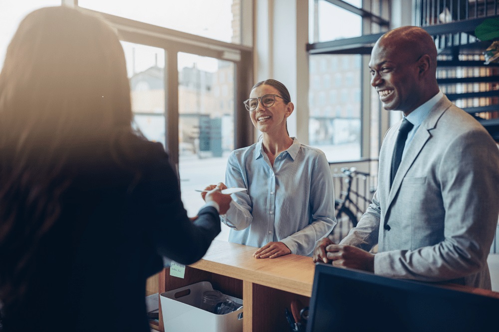 reception-desk-at-hotel Zwei Geschäftsreisende stehen an der Rezeption eines Hotels. Die Empfangsdame gibt ihnen die Unterlagen für die Gruppenbuchung zurück.