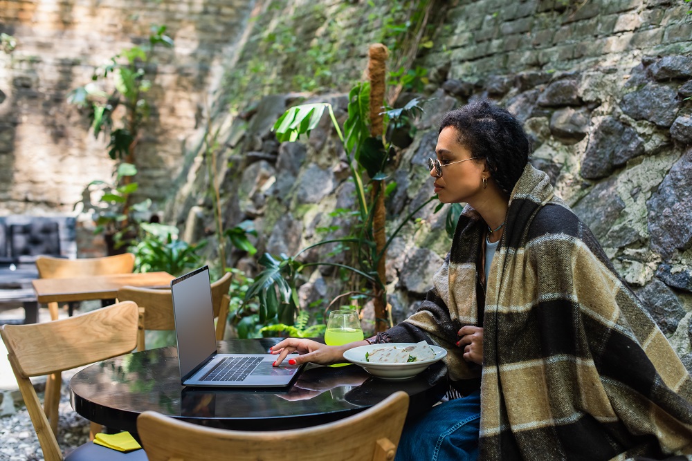 african american freelancer covered in blanket using laptop and having lunch in cafe on terrace Girl working remotely with a stone wall behind her