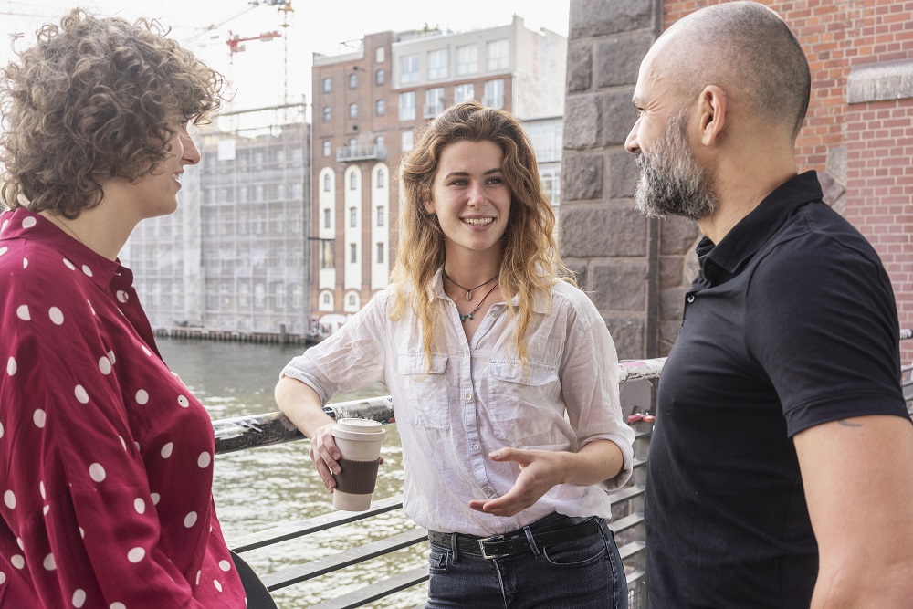Man and female friends talking, river and buildings in background, Berlin, Germany Team members talking on bridge with coffee cup
